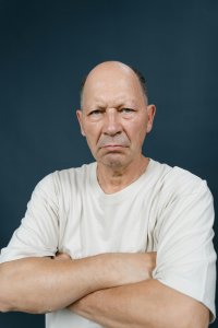Portrait of a bald elderly man with arms crossed showing intense expression in a studio setting.